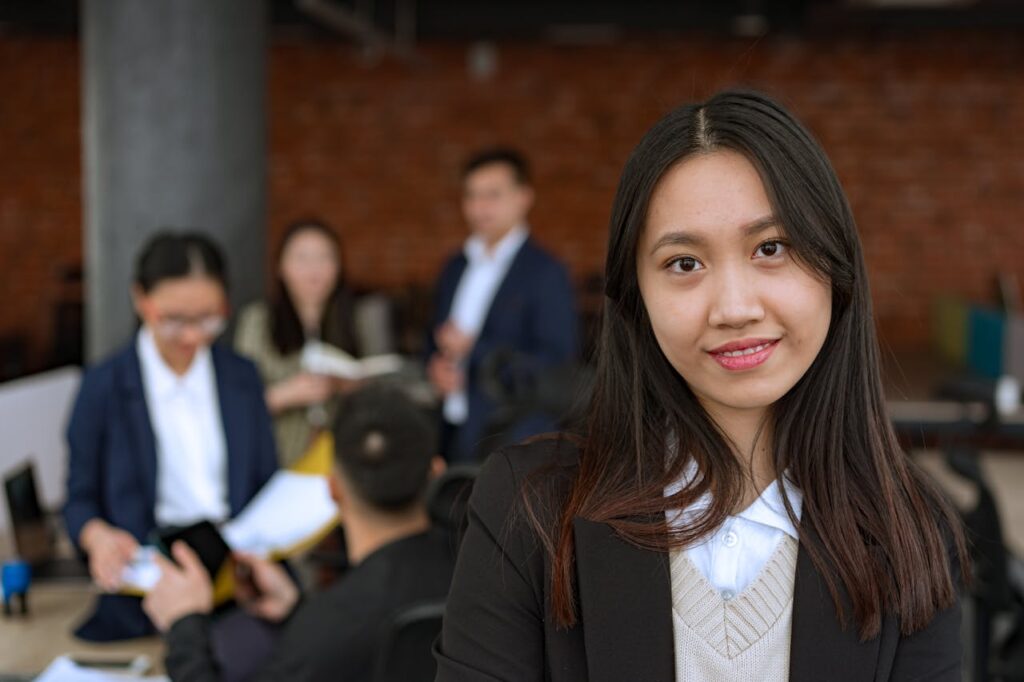 pexels-photo-8101981 Portrait of a confident Asian businesswoman smiling in a modern office with colleagues in the background.